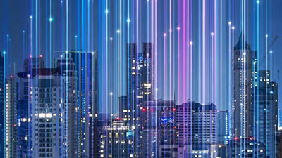 A city skyline at night with brightly lit skyscrapers. Vertical lines of colorful lights, mainly blue and purple, descend from the sky, creating a futuristic and digital atmosphere.