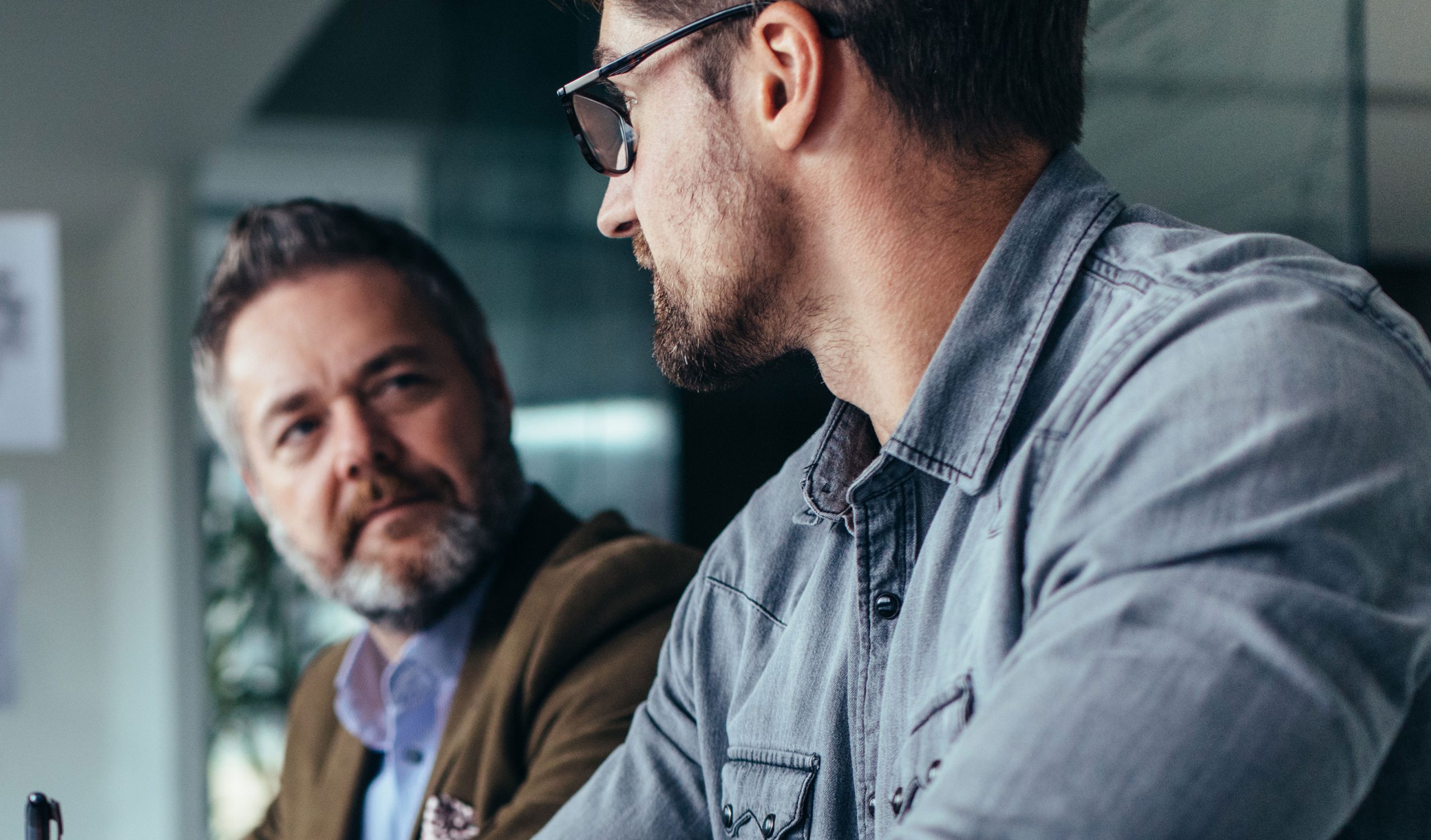 Two men engaged in conversation at a table. The man on the left has gray hair and a beard, wearing a blazer. The man on the right has short hair, a beard, and glasses, wearing a denim shirt. Both appear focused on each other.