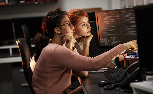 Two women focus intently on a computer screen displaying code in a dimly lit government office. One points at the screen, suggesting collaboration. They are surrounded by telecommunications equipment and shelves filled with binders.