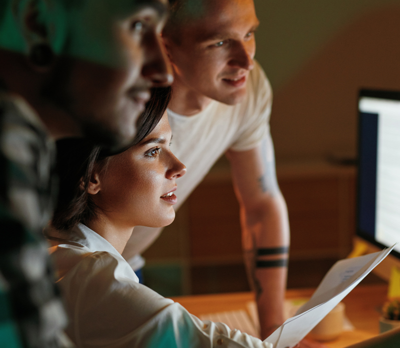 Three people gather around a computer screen, focused and engaged, discussing the Custom 400G from Windstream Wholesale. One holds a document, highlighting their teamwork in an office environment, with warm ambient light illuminating their faces.
