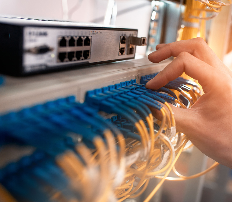 A person adjusts ethernet cables in a network server setup. The close-up view shows a hand interacting with blue and yellow cables connected to a switch, highlighting the precision in maintaining IT infrastructure with custom 400G from Windstream Wholesale.