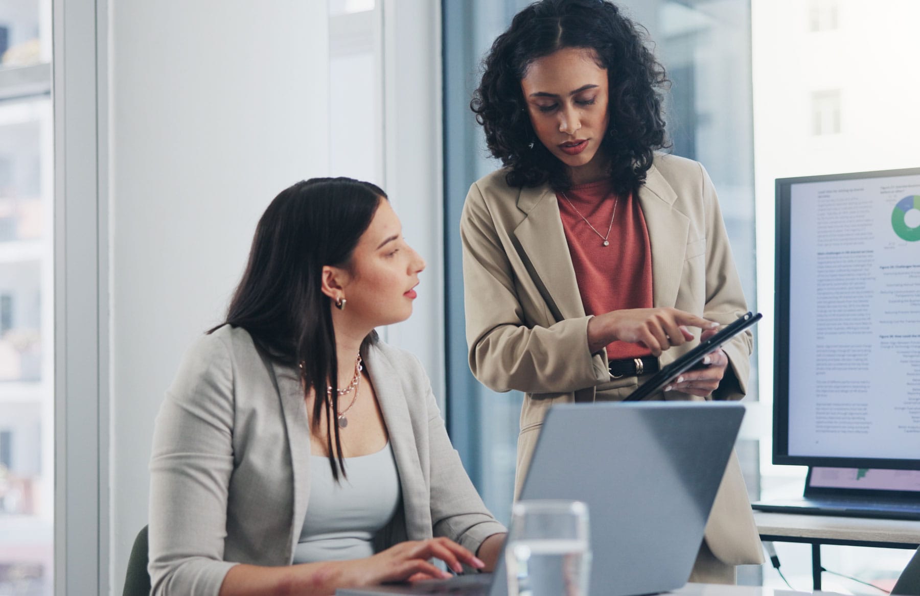 Two women in business attire work in an office; one is seated at a laptop while the other stands beside her, pointing at a tablet. A monitor displaying charts and documents highlights their focus on nationwide network technology.