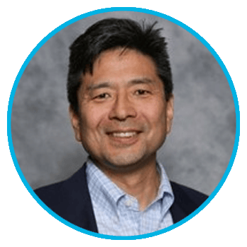 A man with short dark hair smiles at the camera. He is wearing a dark blazer over a light blue checkered shirt, reflecting his customer driven structure. The photo has a gray background and a circular blue border.