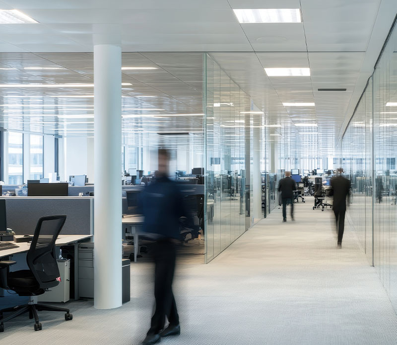 Modern open-plan office with glass walls, rows of desks, and several people walking through the hallway, creating a sense of motion and activity. The environment is bright with ample natural and artificial light.