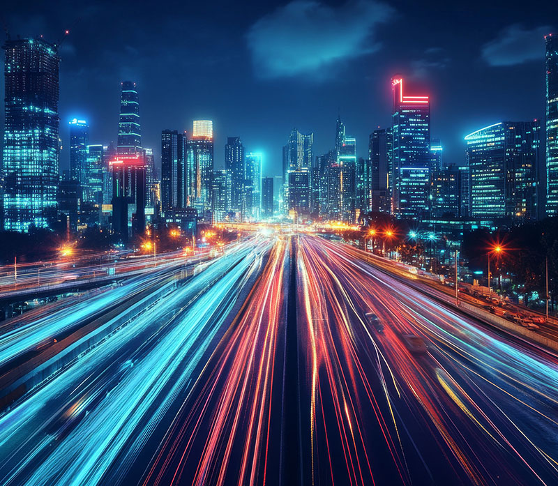 A vibrant cityscape at night features tall, illuminated skyscrapers and bright streaks of car lights on a busy highway, creating a dynamic, futuristic atmosphere under a partly cloudy sky.