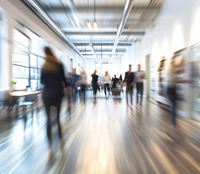 A bright, modern office hallway with people walking and talking, creating a motion blur effect that emphasizes movement and activity in the workspace.