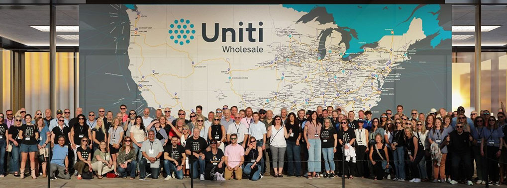 A large group of people pose in front of a map of the United States labeled "Uniti Wholesale." The map covers the entire wall behind them, and the group appears to be at a company event or gathering.