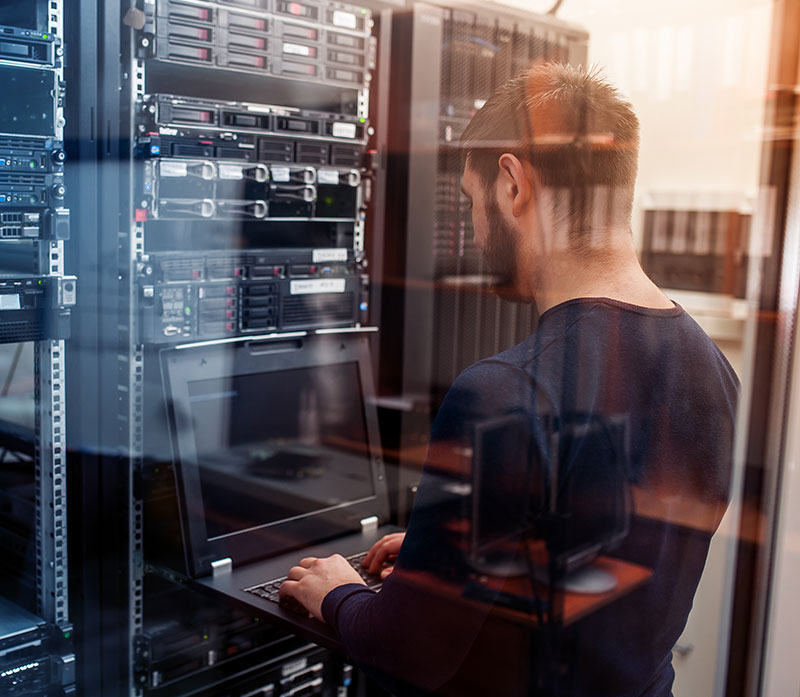 A man stands in a server room using a laptop connected to a server rack. He faces away from the camera, surrounded by computer servers and networking equipment. The scene is reflected on a glass panel.