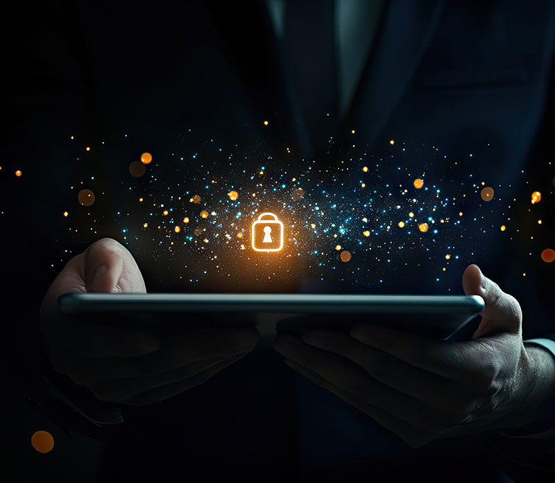 A person in a suit holds a tablet, with a glowing padlock icon and digital particles hovering above the device, symbolizing cybersecurity and data protection.
